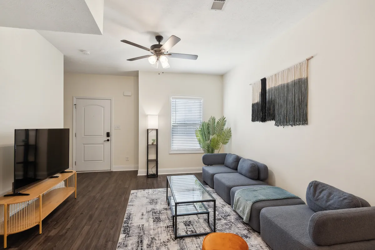 A cozy living room with plush seating arranged around a modern coffee table, a large television mounted on the wall, stylish wall decor, a floor lamp, and a healthy green plant in the corner. The space is bathed in warm lighting, inviting both relaxation and social gatherings.