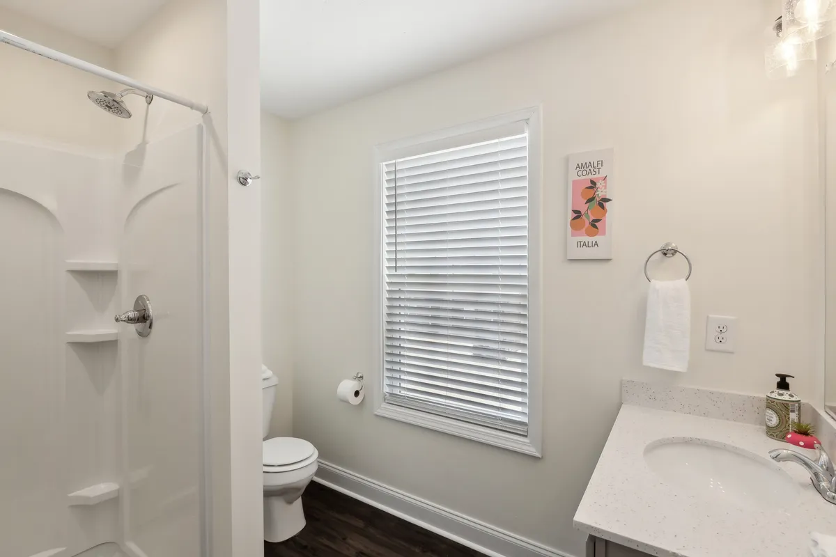 This image features a full bathroom with a shower area, sink with mirror, toilet, window with blinds, and wall artwork. The layout emphasizes both function and style, including personal touches that make the space feel inviting and well-maintained.
