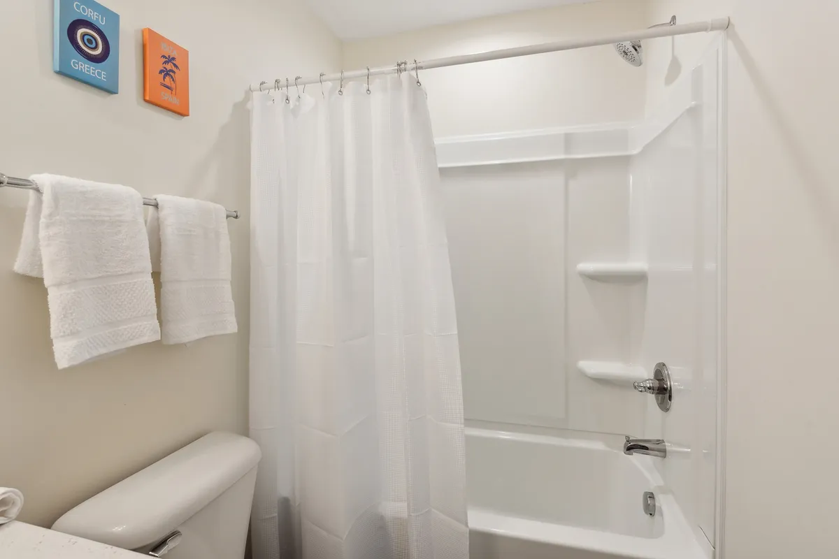 A serene bathroom scene showing a white shower curtain, colorful wall art, a neatly stacked set of white towels, toilet, and bathtub. The space evokes relaxation and cleanliness, with small details adding a cozy, welcoming feel.