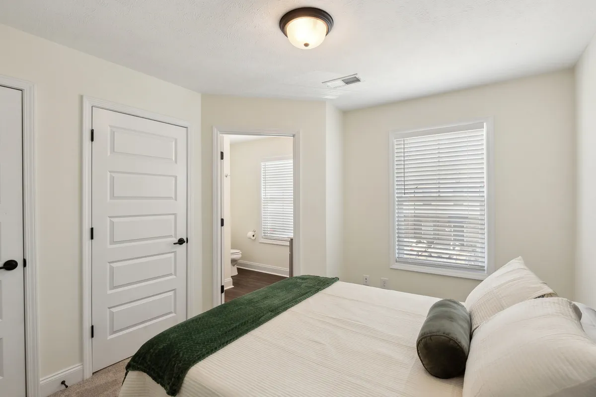 This image showcases a serene bedroom with a cozy bed, a green throw blanket, and sunlit windows. The tranquil setting suggests a comfortable, restful space where guests can unwind after a day of exploring Clarksville with friends or colleagues.