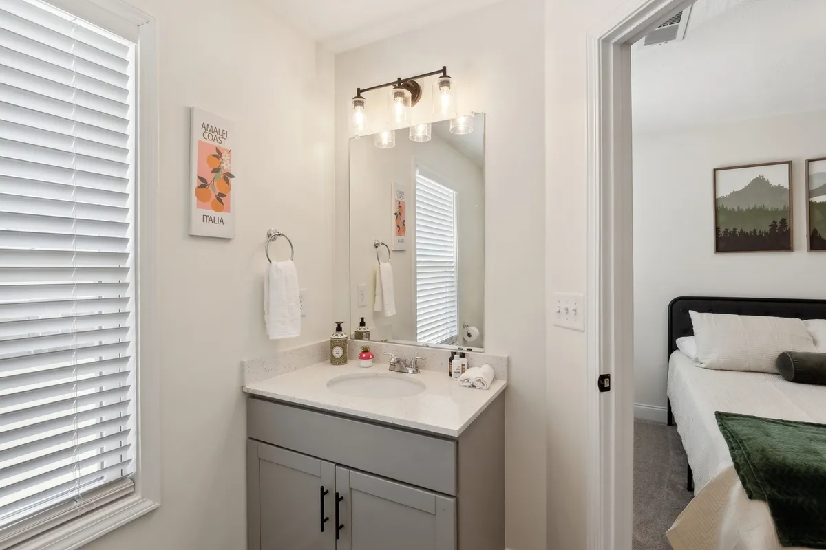 A stylish vanity area with a large mirror, sink, and contemporary artwork, with a glimpse of a bed in the background. The fresh and airy ambiance highlights the apartment's well-appointed amenities and the comfort it offers for preparing before heading out or relaxing with friends.