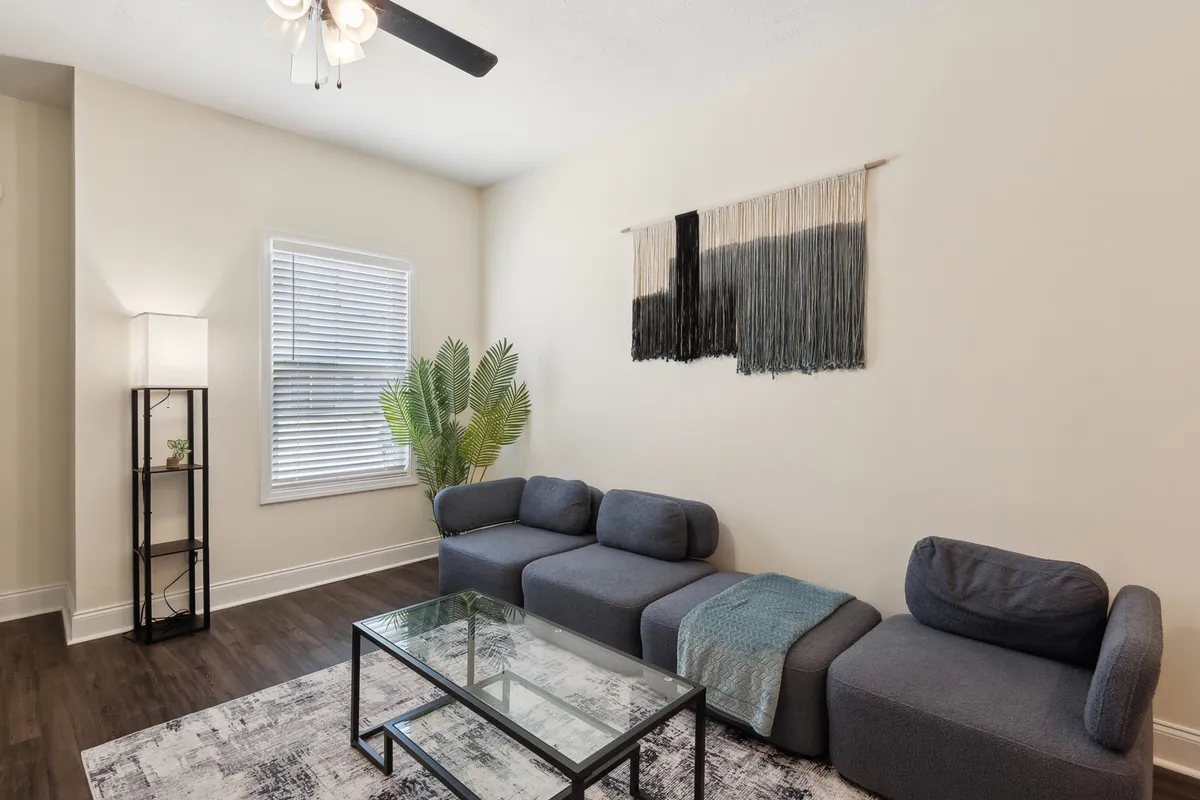 A modern living room with a plush sofa, glass coffee table, stylish wall hanging, a touch of green from a potted plant, and a warm lamp. Natural light pours in from a window, creating a welcoming atmosphere for both relaxation and brief meetings.