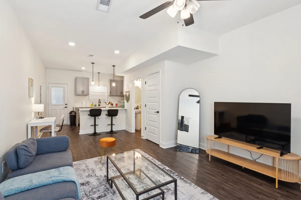 A contemporary open-plan space with a cozy sofa facing a modern TV, a sleek coffee table, a kitchen bar with seating, and a large mirror that enhances the sense of space. The setup is perfect for relaxing, watching TV, or entertaining guests, blending comfort with modern style.