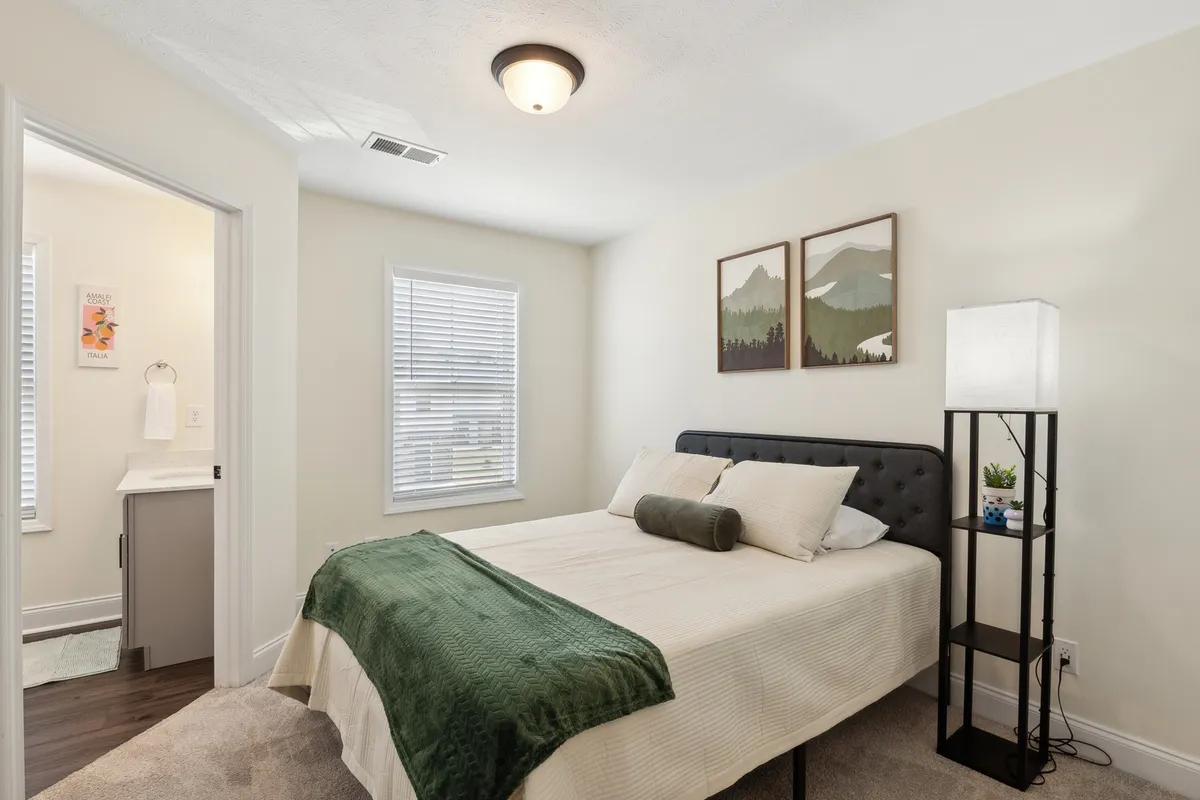 A serene bedroom featuring a neatly made bed with plush bedding, soft lighting from a bedside lamp, and decorative art on the wall. The room is bathed in natural light, creating a peaceful atmosphere ideal for relaxation or reading.